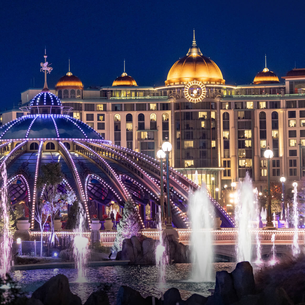 Atlantic restaurant and fountains illuminated at night with Universal Helios Grand Hotel in the background in Celestial Park at Universal Epic Universe.