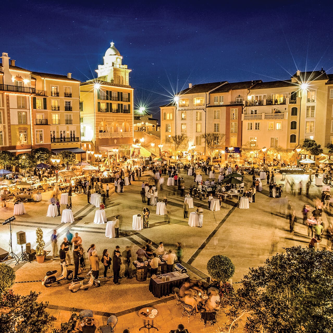 An exterior view of guests enjoying the Harbor Piazza at night with a backdrop of the Loews Portofino Bay Hotel