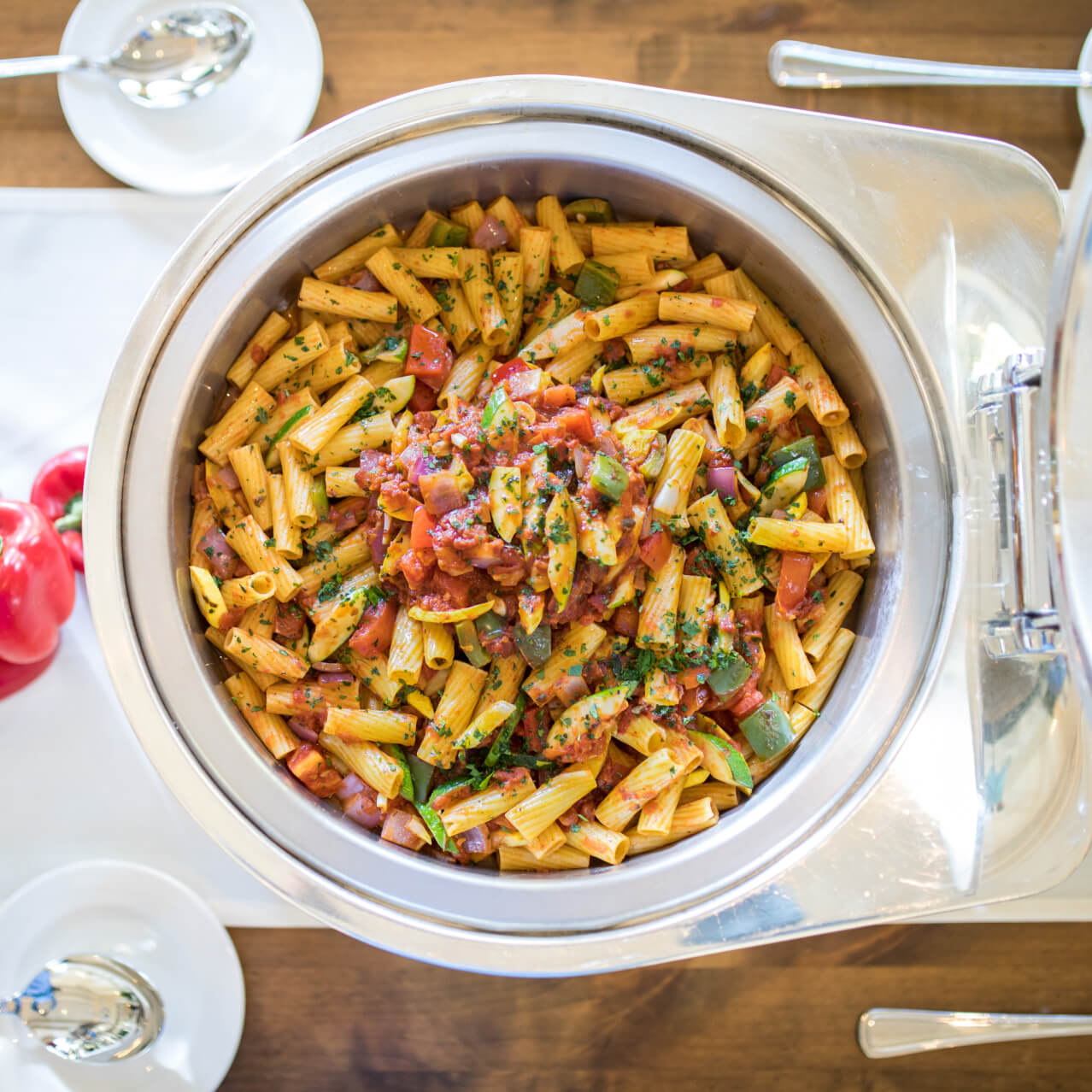 A large chafing dish on a buffet table featuring pasta with tomatoes and peppers.