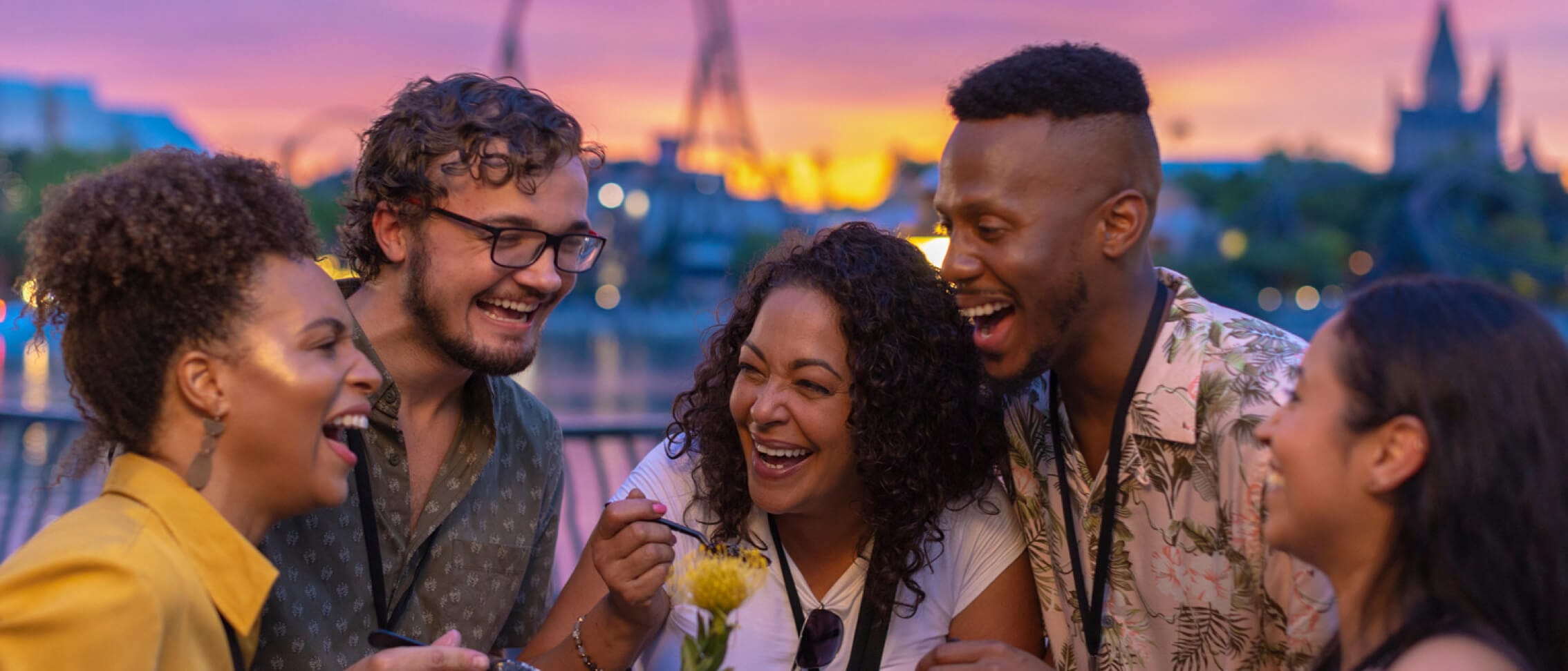 A group of friends enjoying food in beverages in front of the Jurassic World VelociCoaster roller coaster during sunset.