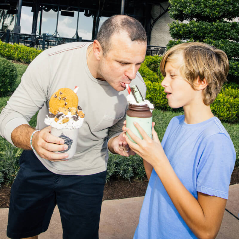 Dad holding a milkshake tasting the milkshake in his son's hand in front of The Toothsome Chocolate Emporium & Savoury Feast Kitchen