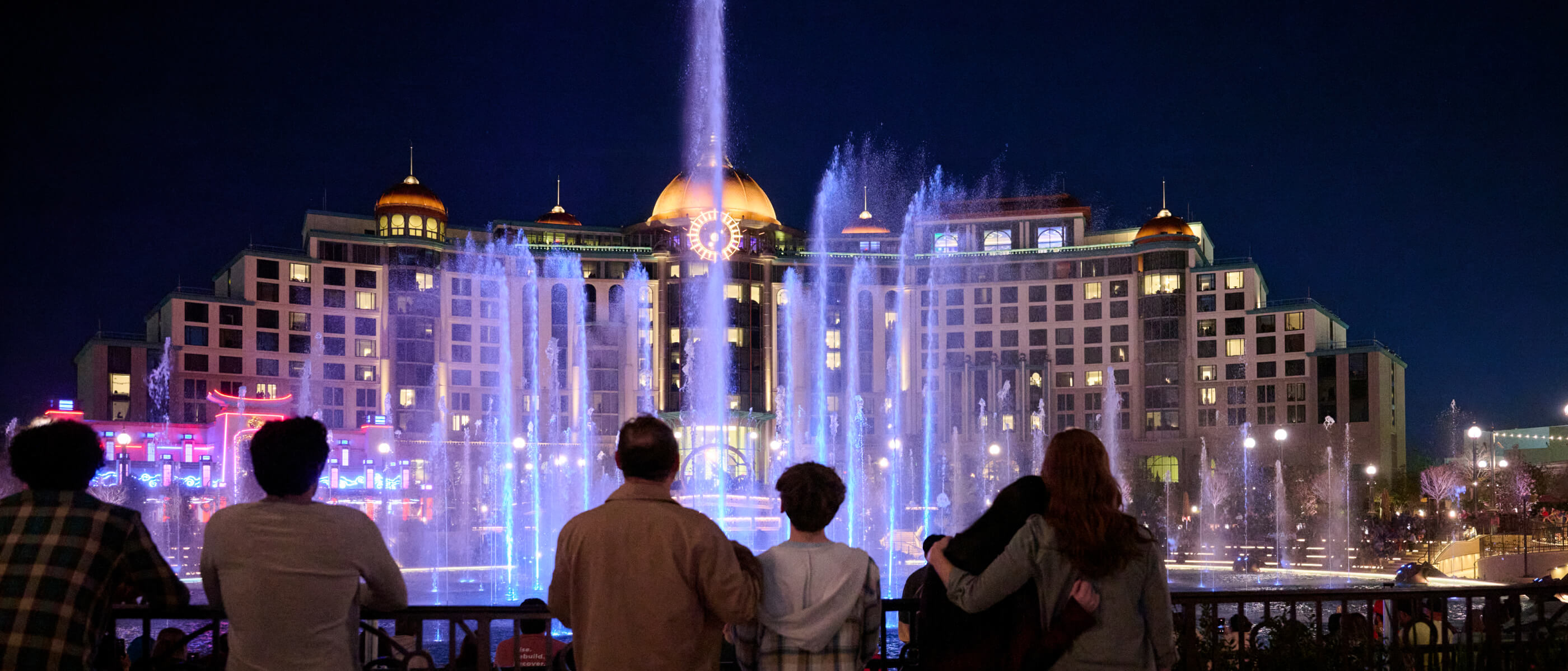 Guests watching from behind a railing as a colored fountains soar into the nighttime air in front of a grand hotel.