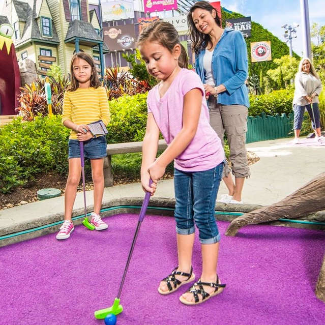 A young girl holding a golf club prepares to hit a ball while her mother and sister look on at Hollywood Drive In Golf at Universal CityWalk in Orlando.