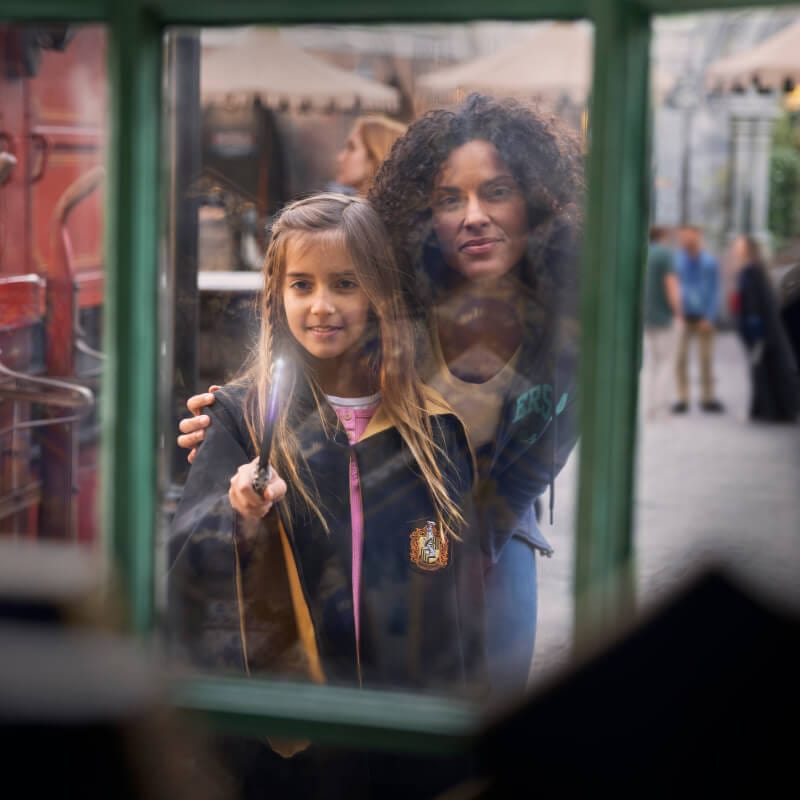 A child in wizard robes holding an illuminated wand up to a window while her mother looks on.