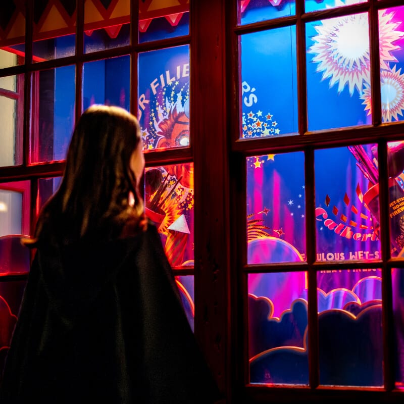 A guest in wizard robes looking at a colorful window display in The Wizarding World of Harry Potter™.