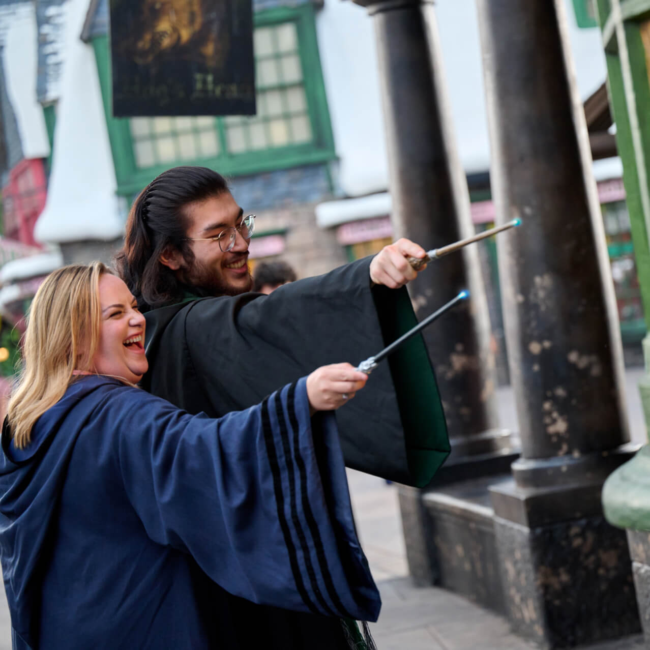 A pair of excited Guests in wizarding robes pointing their glowing wands at a storefront window.