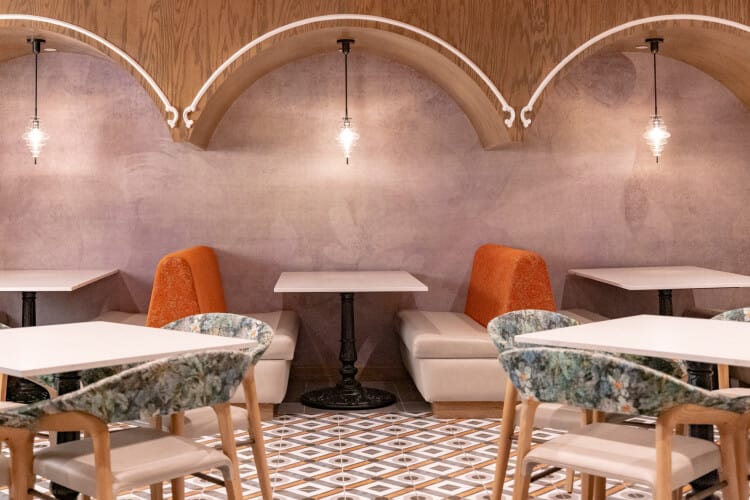 A warmly lit lounge area with recessed booths in the background and square tables with chairs in the foreground.