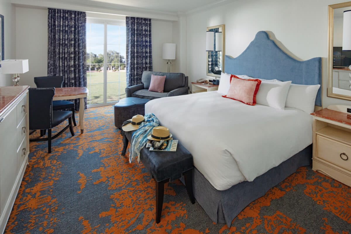 Portofino Bay Hotel standard room with king bed and side tables, large dresser and a corner table plus large stuffed chair and ottoman beside large window in background.