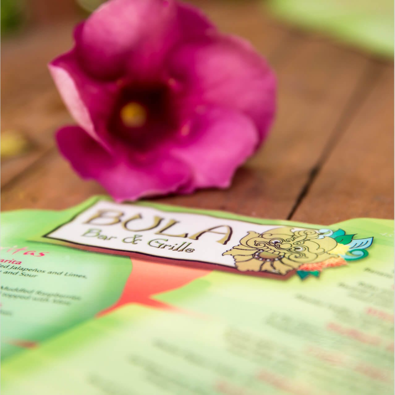 A Bula Bar & Grille menu sits on a table beside a vibrant pink flower.