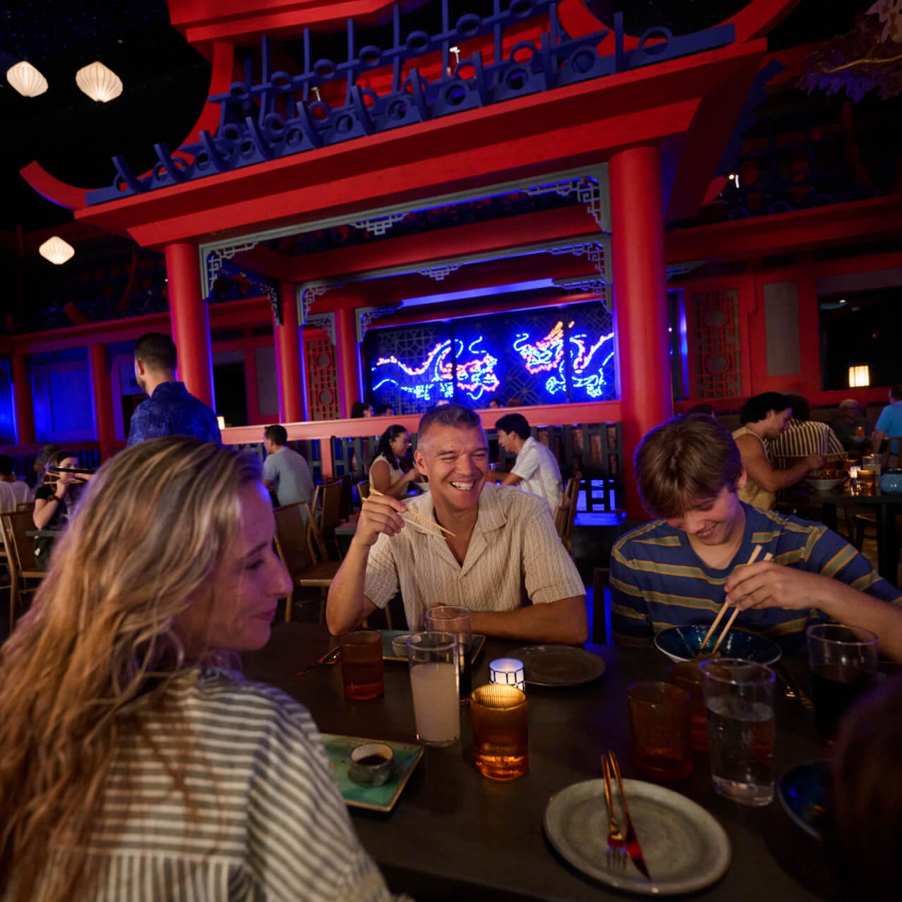 A smiling family eating food with chopsticks in a themed restaurant setting with red archways and neon dragon decorations.
