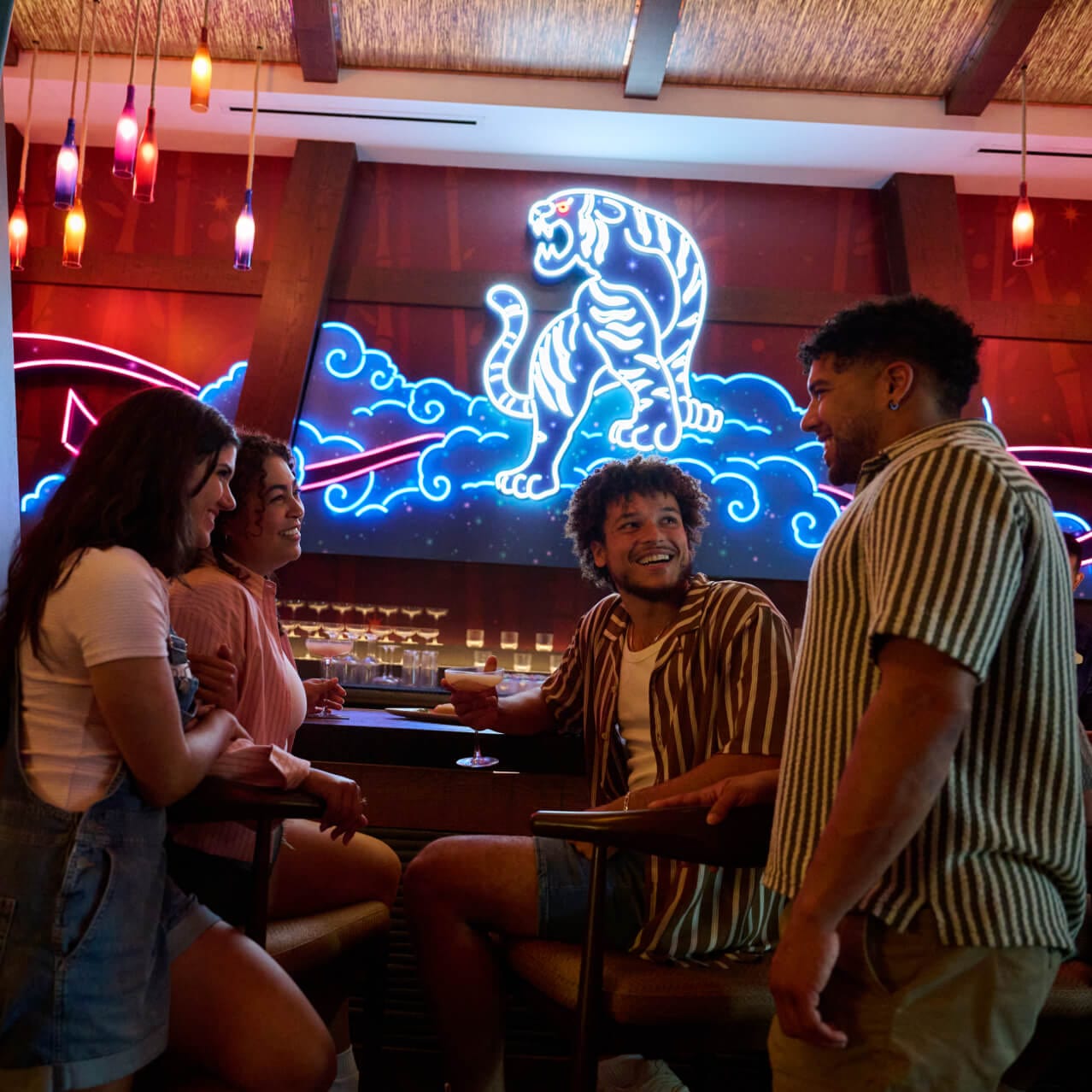 A group of smiling friends enjoying drinks in a bar setting with a striking neon tiger design on the wall behind them.