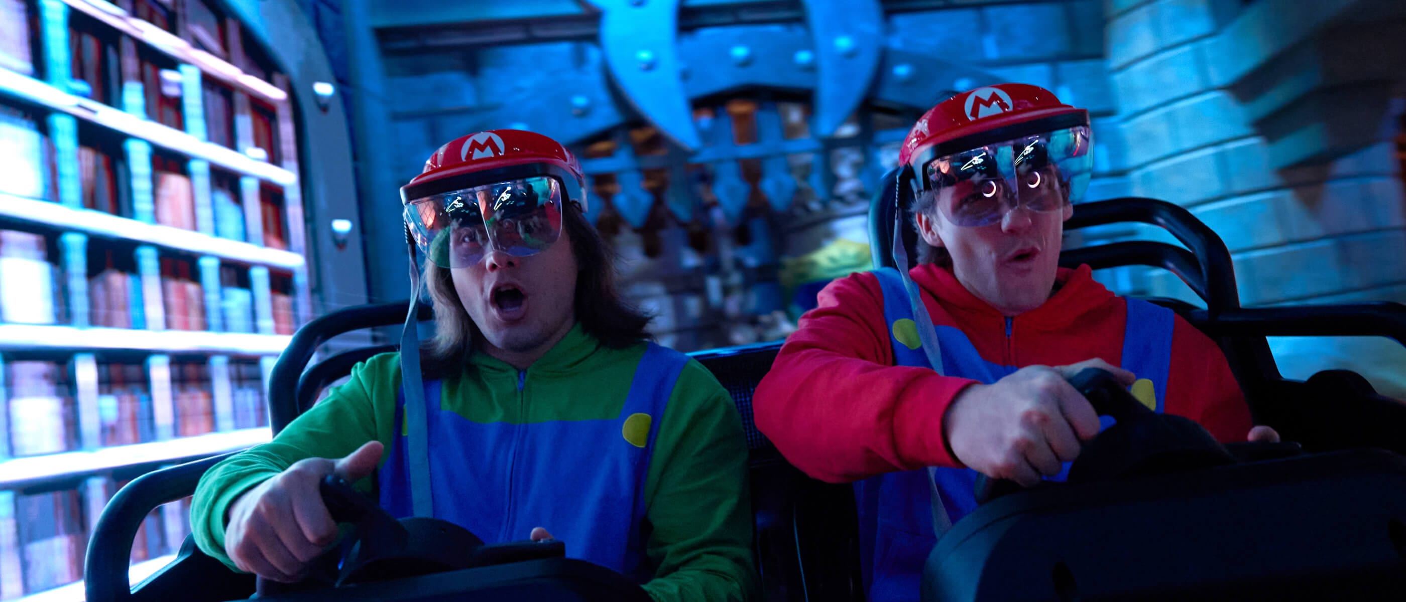Two friends in Mario and Luigi shirts and virtual goggles turning their steering wheels with a castle wall in the background.