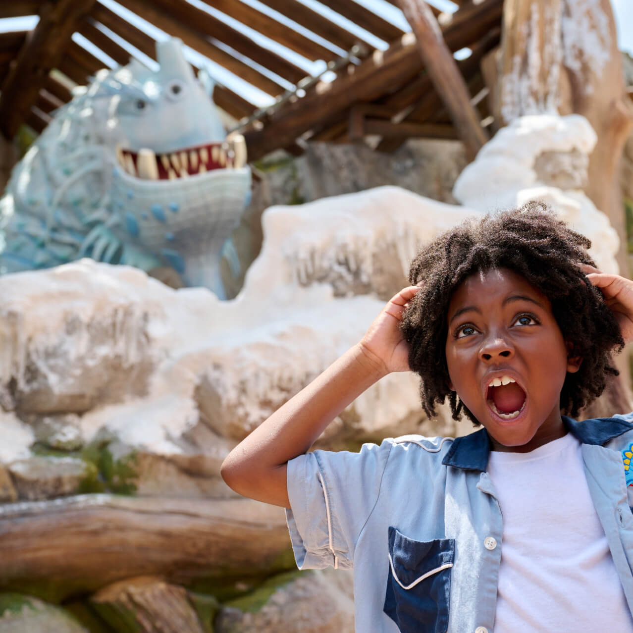 A child screams in delight with his hands on his head as a large, light blue dragon appears above the rocks behind him.