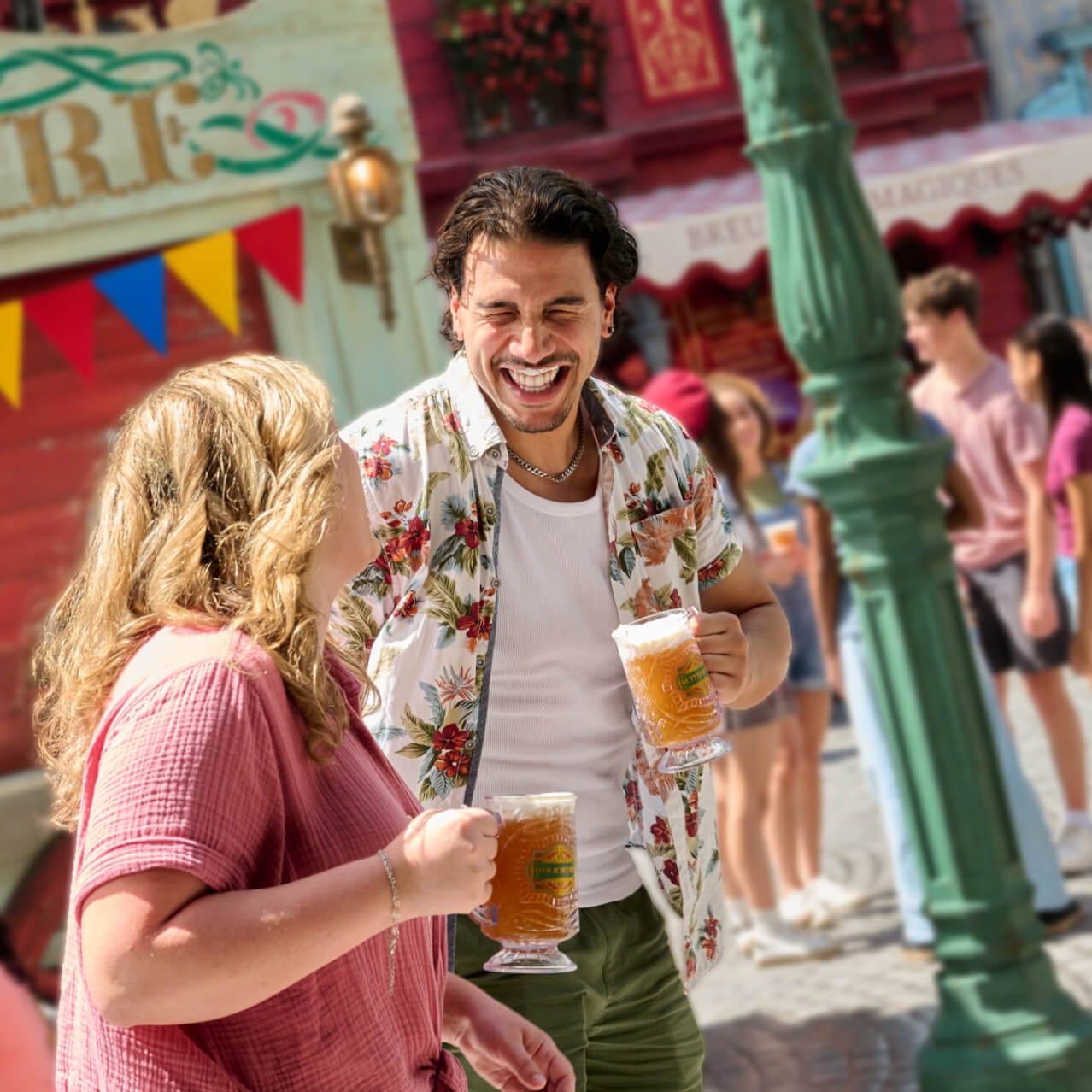 A couple laughing while holding mugs of Butterbeer™ and strolling through the colourful streets of wizarding Paris.