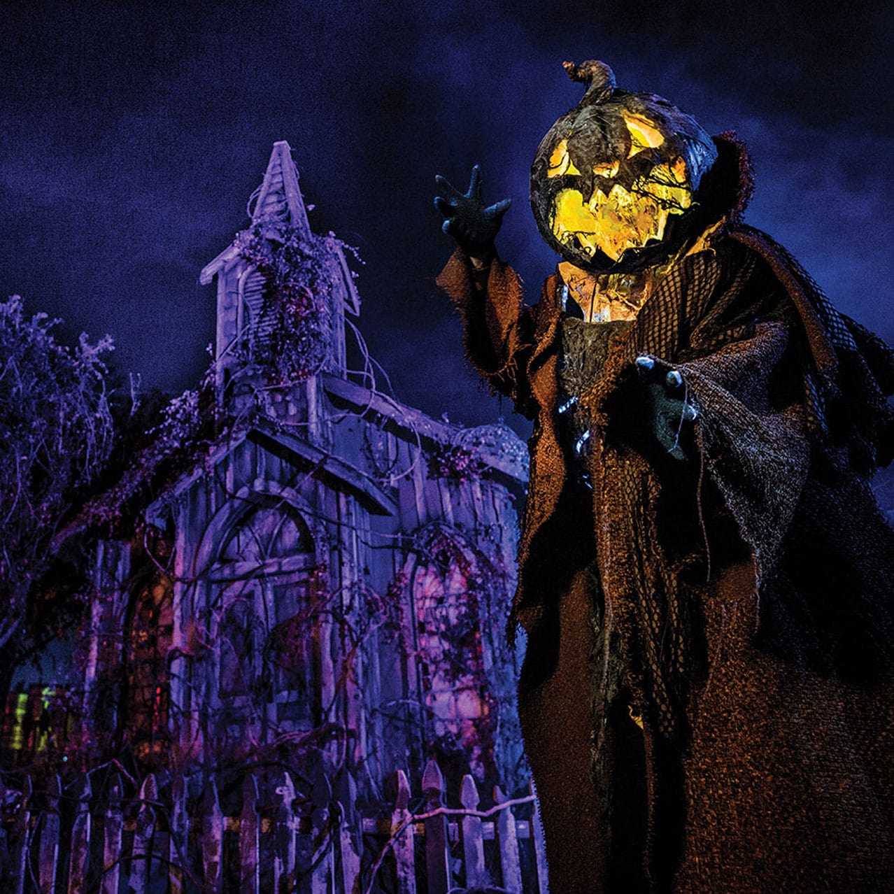 A creepy pumpkin-headed figure beckons towards a vine-covered building at Halloween Horror Nights.