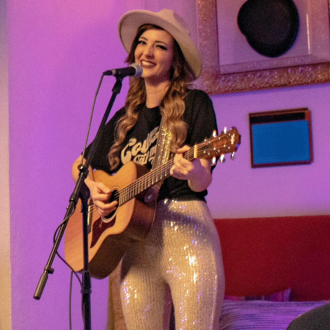 A woman with an acoustic guitar stands at a mic and performs live music for a small crowd inside the Velvet Bar.