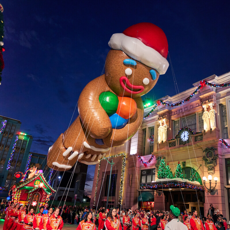A large float balloon of the Gingerbread Man in a Santa hat holding colorful candies passing festively decorated buildings.