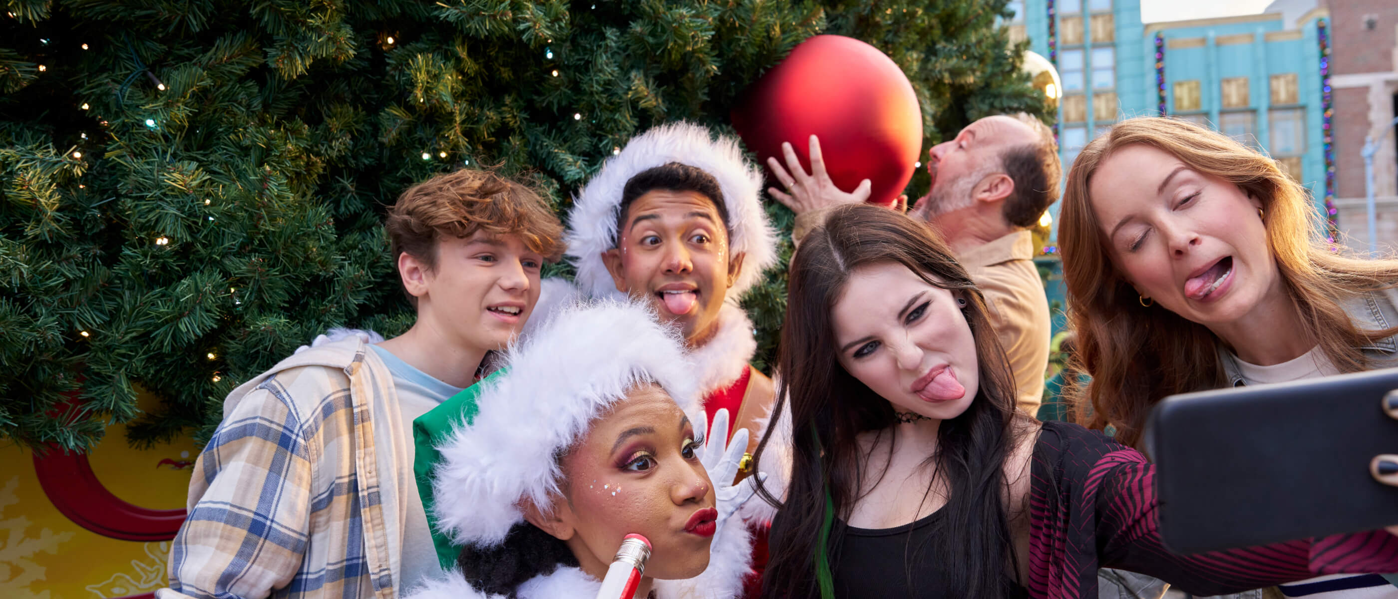 A family posing for a selfie and making silly faces with festively dressed Whos in front of a giant Christmas tree.