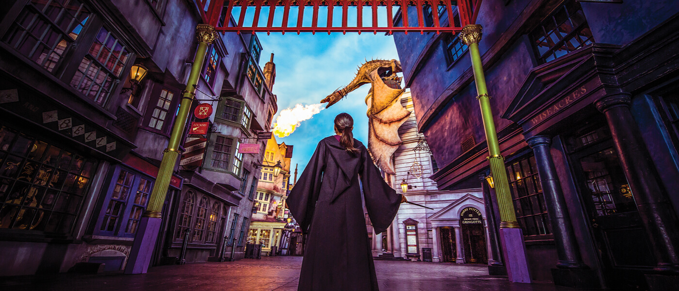 A girl in a wizard robe stands alone in The Wizarding World of Harry Potter™ - Diagon Alley™, as the dragon blows fire.