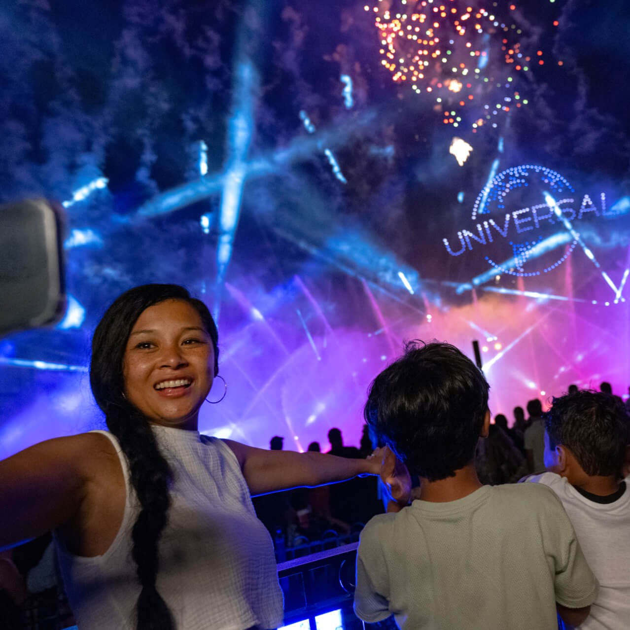 A Guest taking a selfie as her family watches a nighttime show with fountains, fireworks and the “UNIVERSAL” globe in lights.