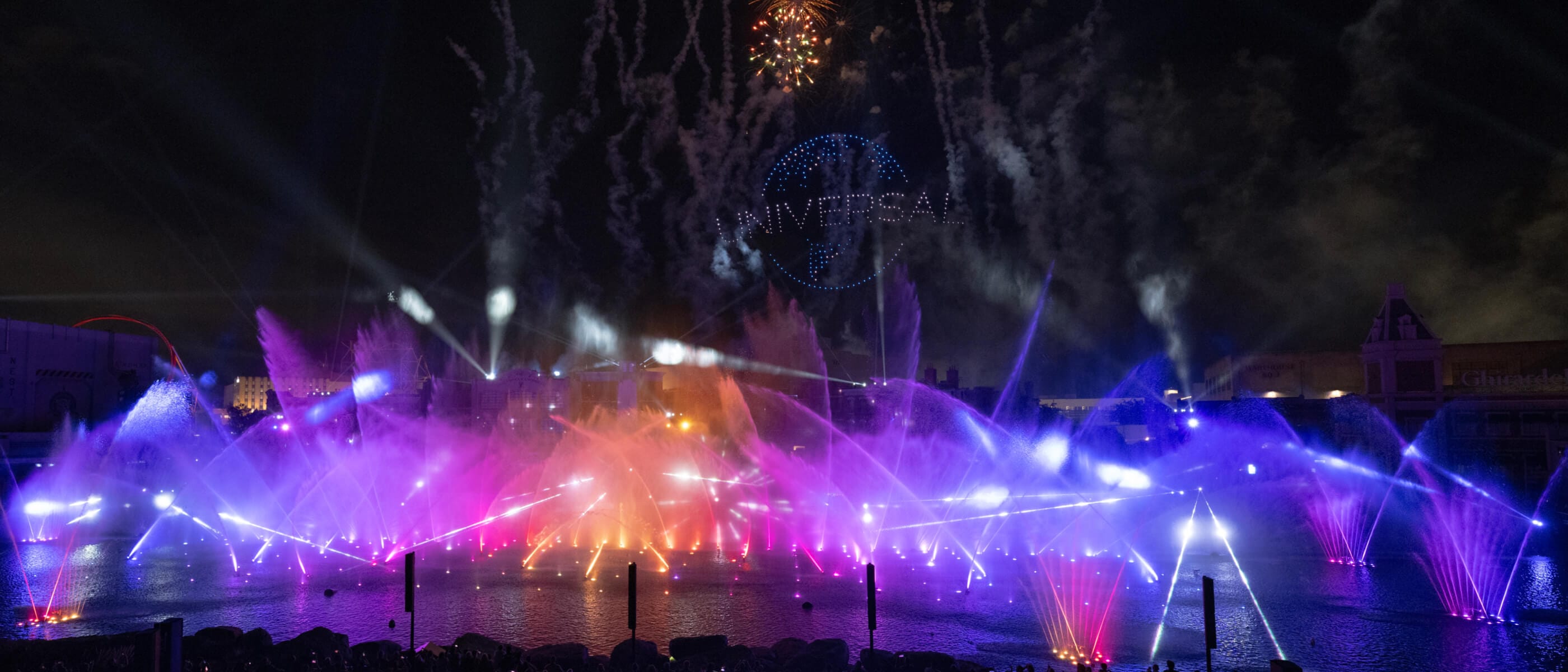 A nighttime show with multicoloured fountains on the water and fireworks and the "UNIVERSAL" globe in lights in the sky.