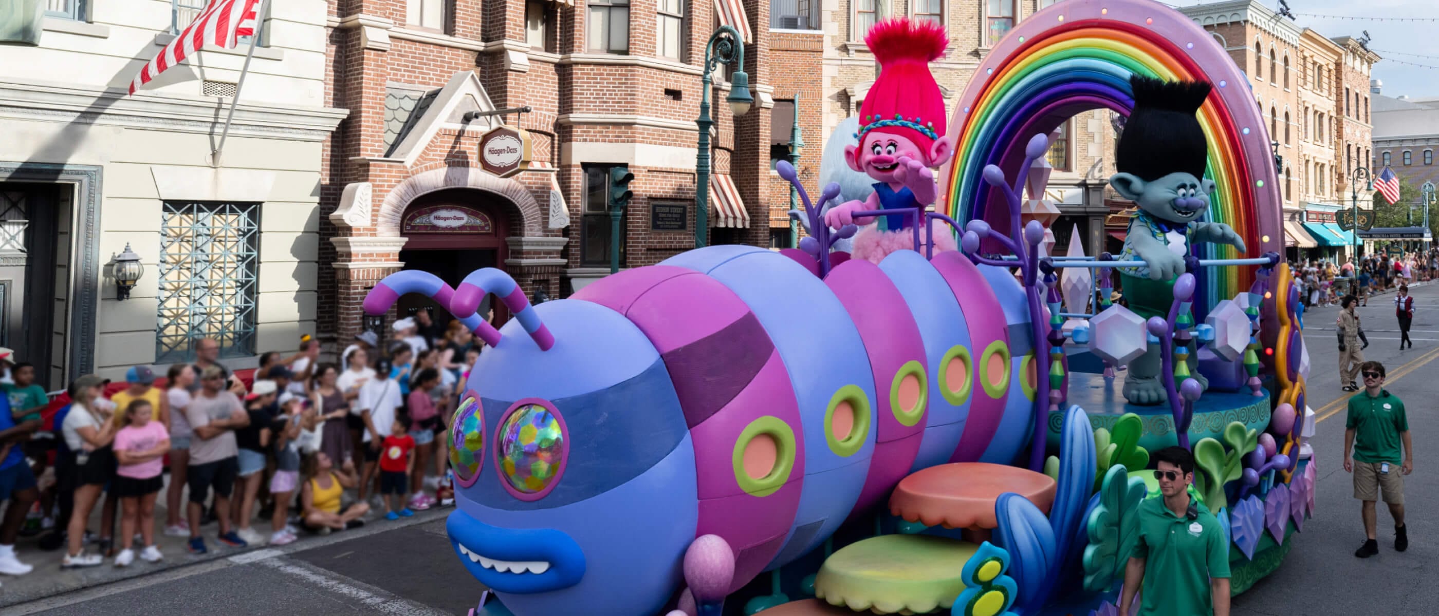 Trolls film characters riding on a colourful parade float shaped like a caterpillar bus as Guests watch from the pavement.