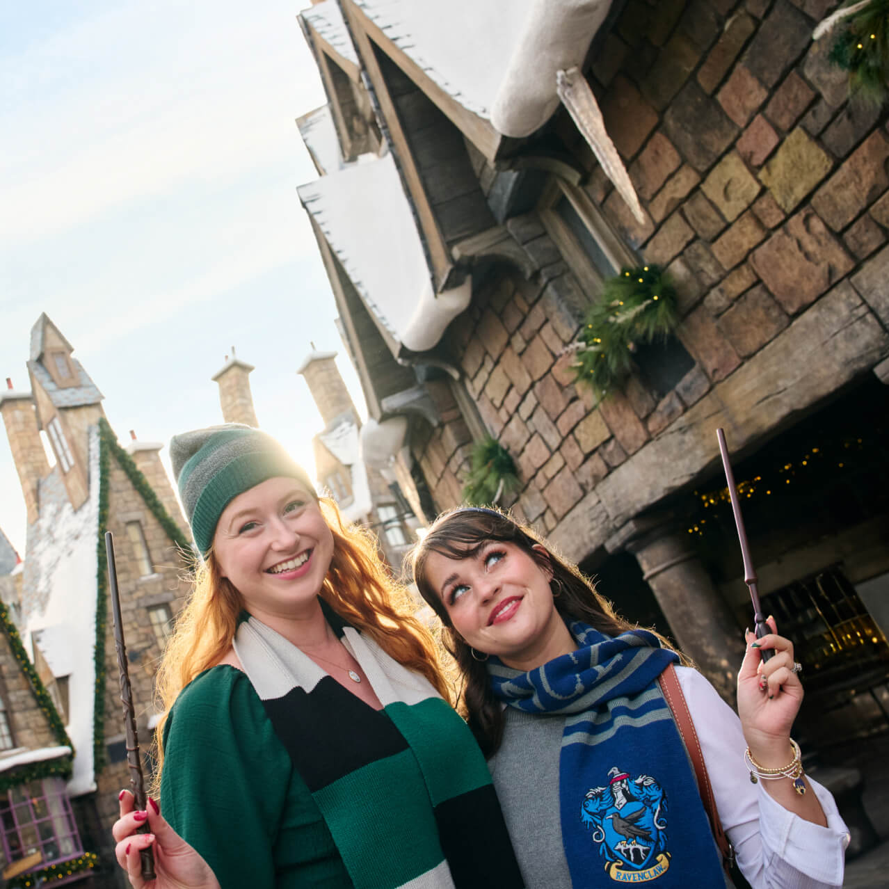 Two friends with Hogwarts scarves and wands amid snowy holiday-decorated buildings in The Wizarding World of Harry Potter.
