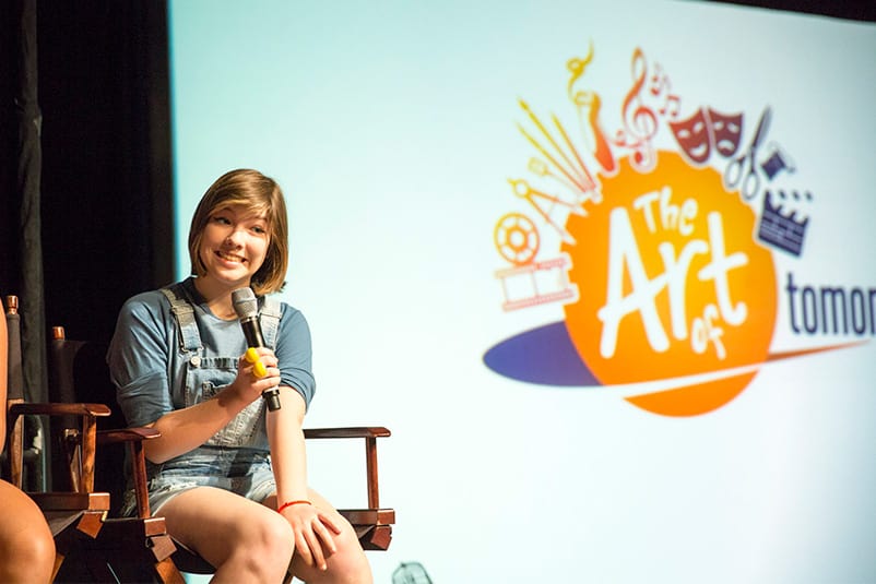Girl sitting on a chair, holding a microphone in front of The Art of Tomorrow screen.