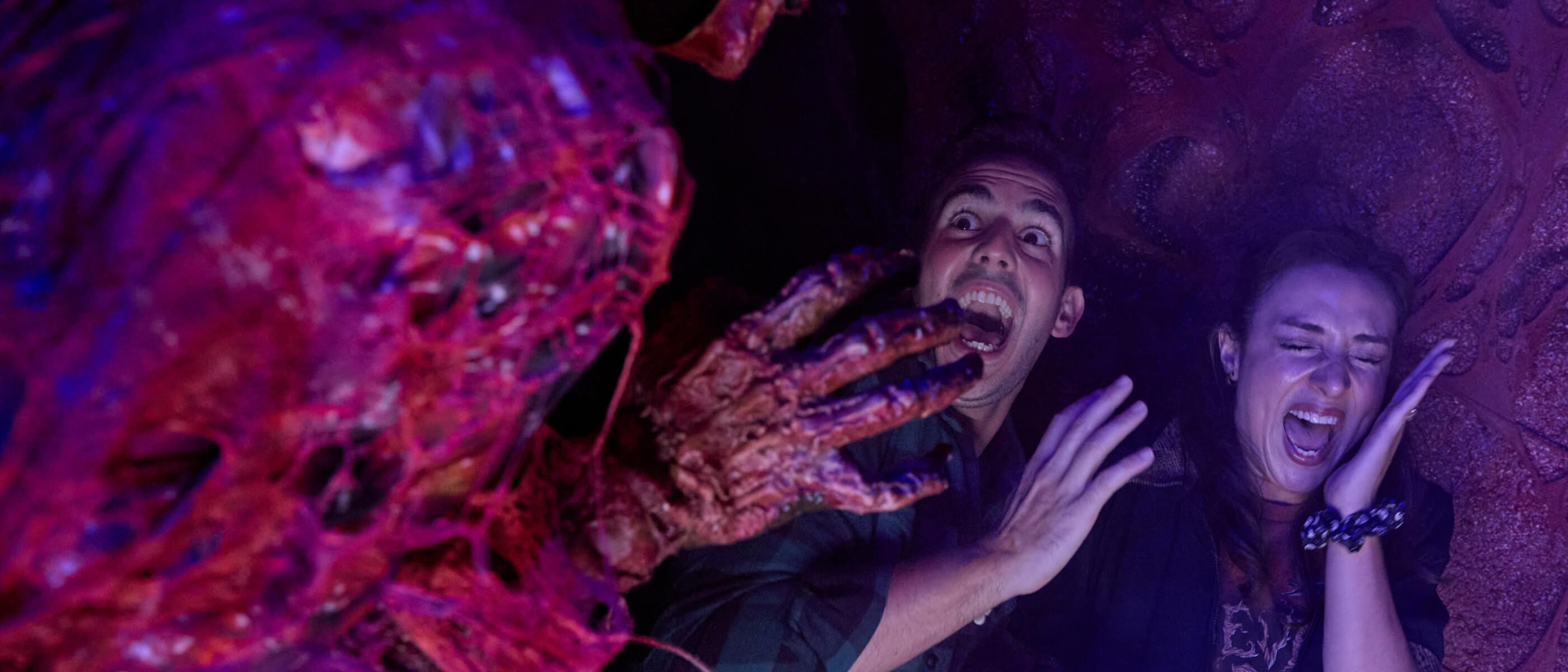 A couple holding hands and screaming in terror as a slimy creature reaches its decaying hand out towards them in the darkness.