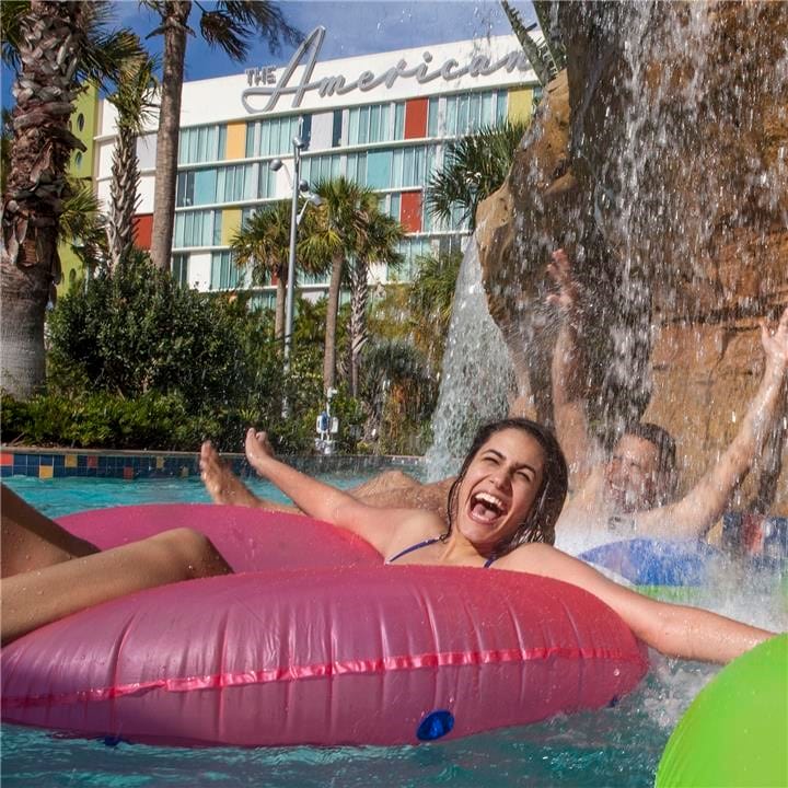 Universal Orlando's Cabana Bay Beach Resort features two family friendly swimming pools and a lazy river that kids and adults alike will love. A woman in a pink inner tube smiles and laughs along with a group of other young adults as they pass a waterfall in the lazy river at Universal's Cabana Bay Beach Resort Orlando.