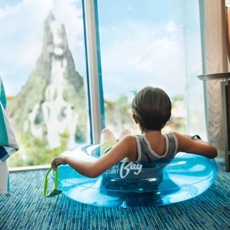 Boy sits in an inner tube in his room at Universal's Cabana Bay Beach Resort while enjoying a view of Universal's Volcano Bay through the window.