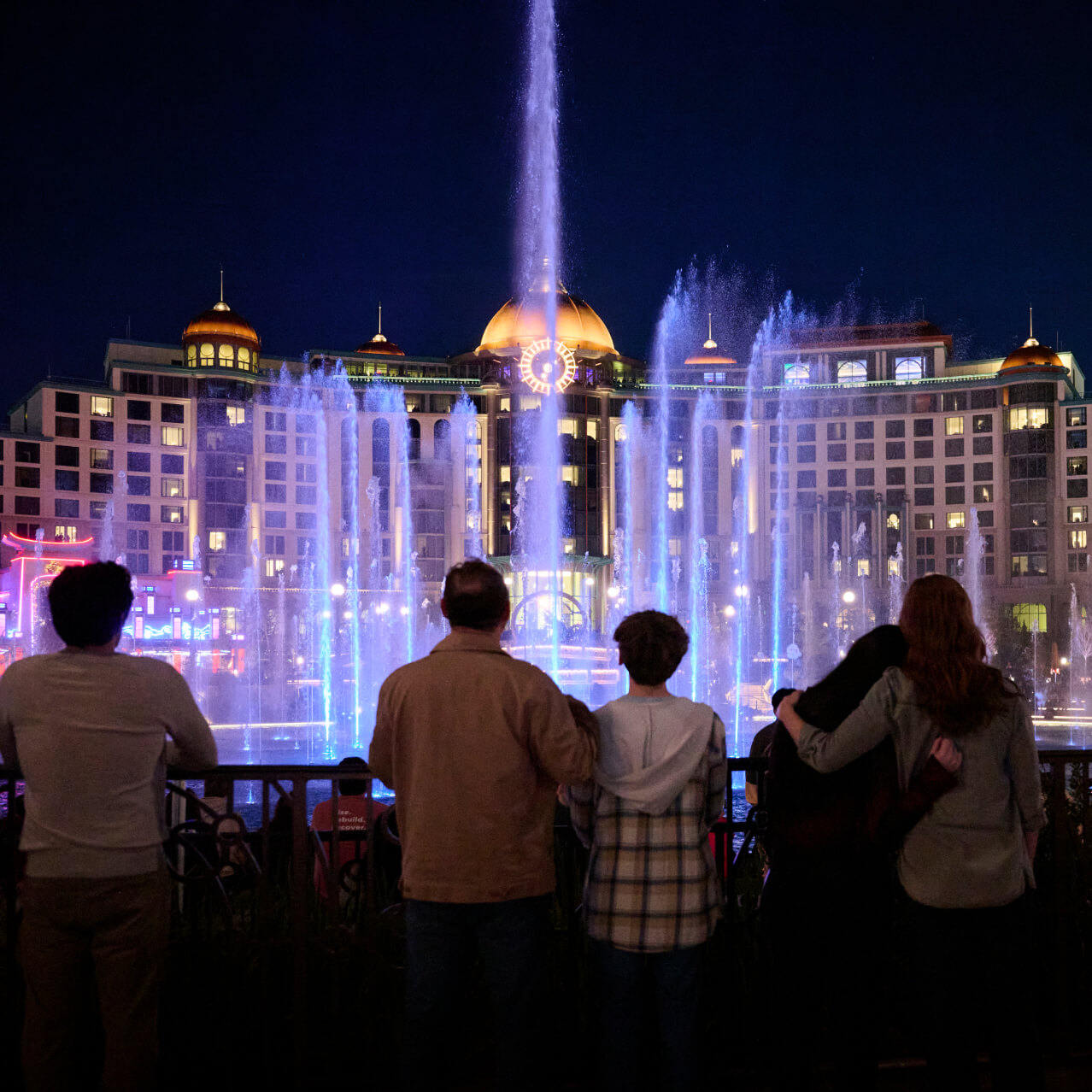 Guests watching from behind a railing as a colored fountains soar into the nighttime air in front of a grand hotel.