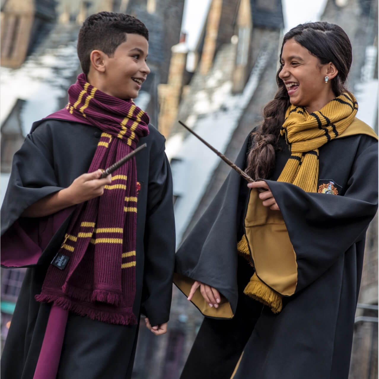 Two children wearing Hogwarts cloaks walking through the streets of a snow-capped Hogsmeade.