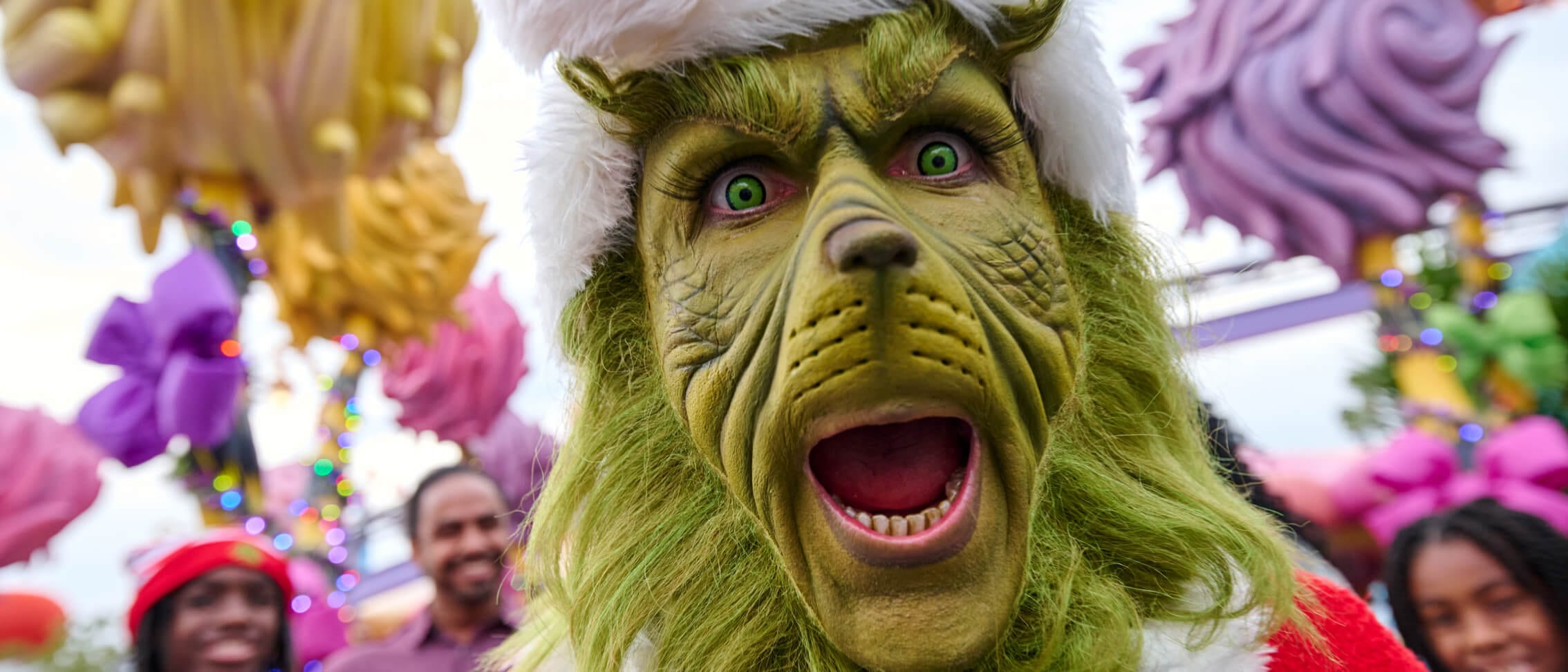 The Grinch dressed as Santa making an excited expression as people stand in the background.