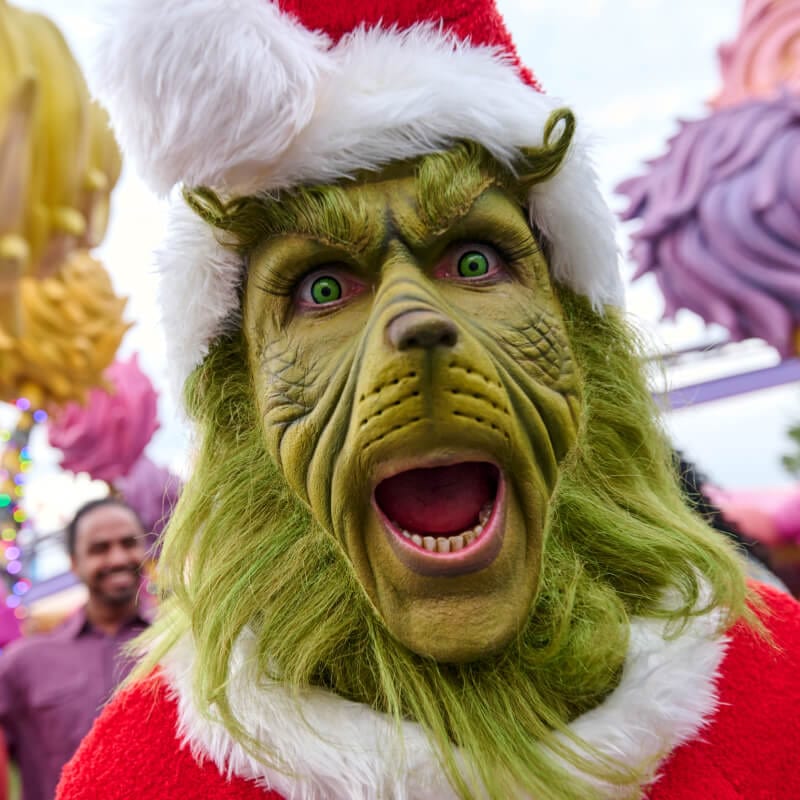 The Grinch dressed as Santa making an excited expression as people stand in the background.
