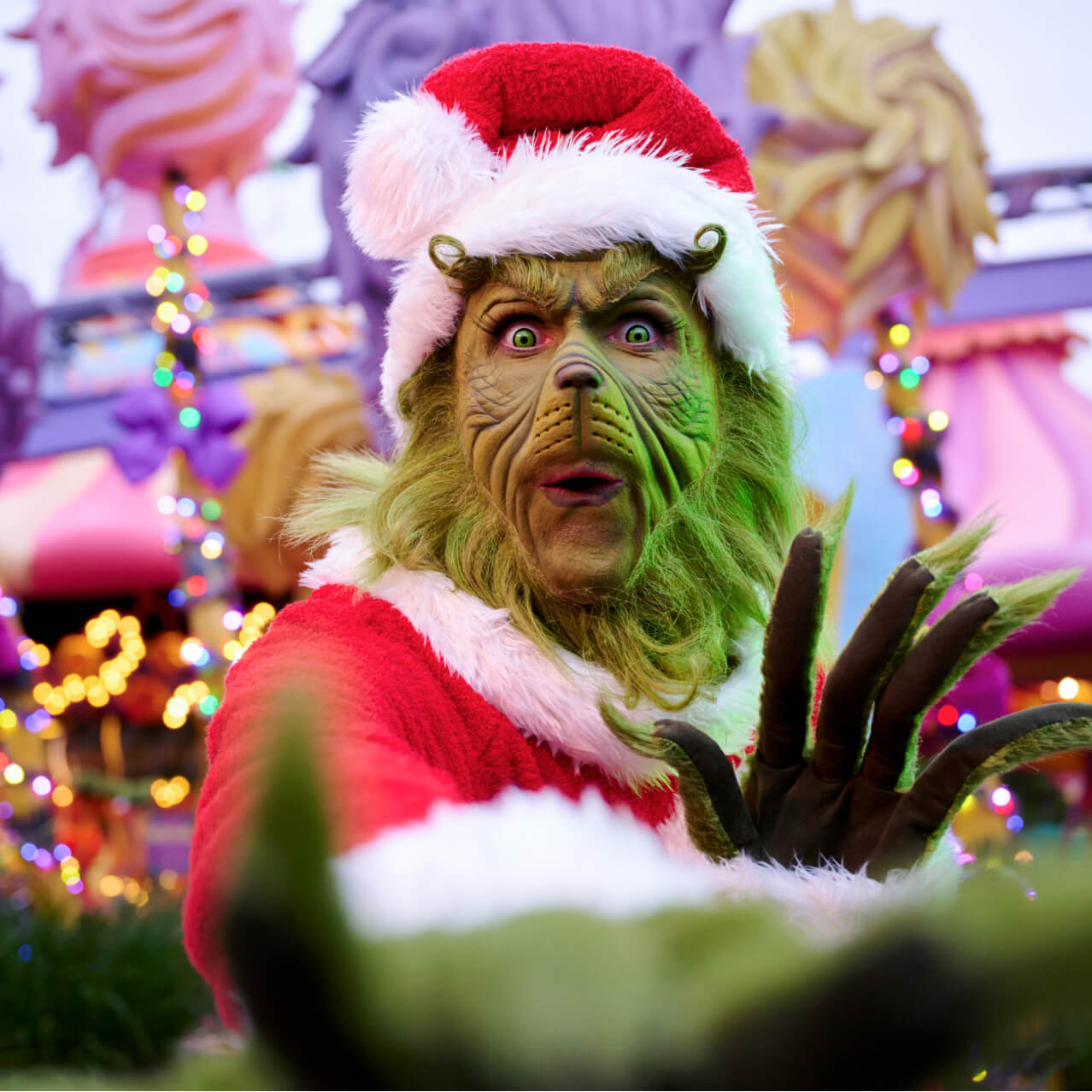The Grinch dressed up as Santa and making a mischievous facial expression with Seuss Landing decorated with Christmas lights in the background.
