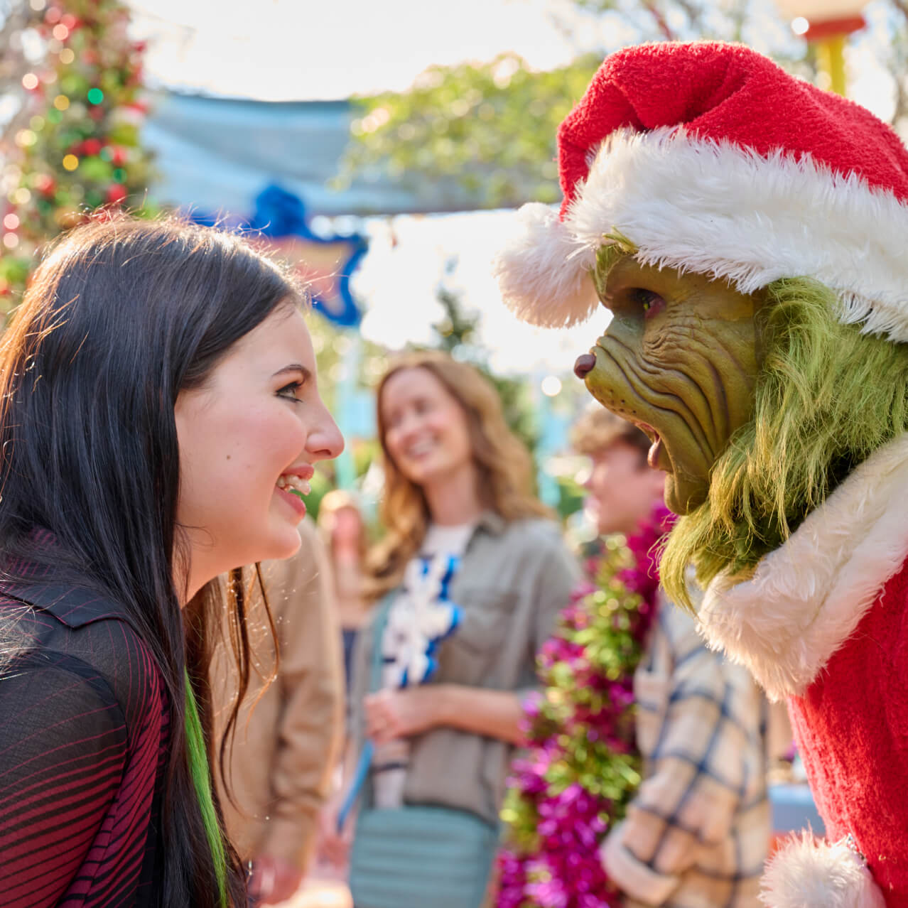 A smiling Guest facing down The Grinch in his Santa hat and coat with other Guests and festive décor in the background.