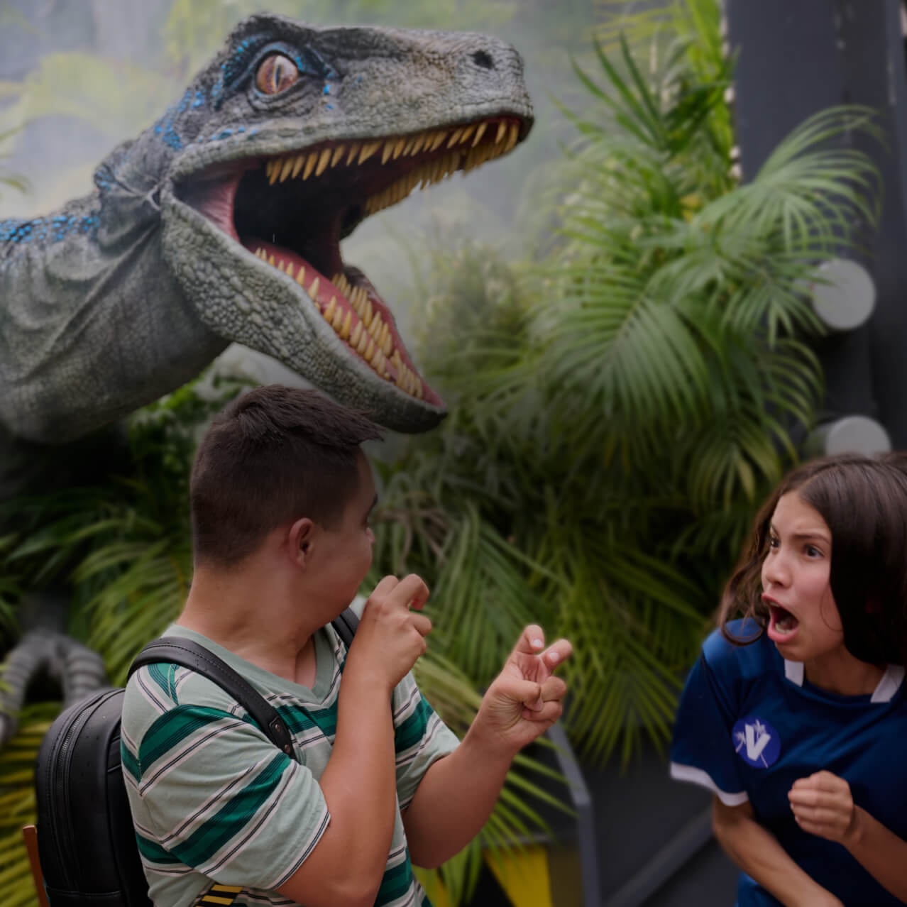 One kid making claws with his hands while others scream as Blue the velociraptor hovers overhead with its mouth open wide.