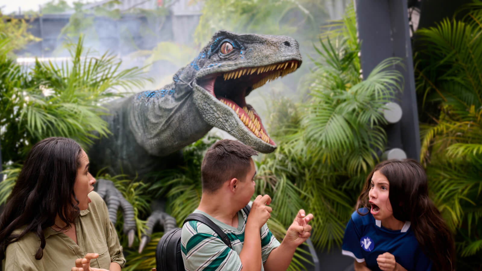 One kid making claws with his hands while others scream as Blue the velociraptor hovers overhead with its mouth open wide.