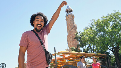 A man at the entrance to Universal Islands of Adventure holding his arm up and pretending to touch Pharos Lighthouse in the background.