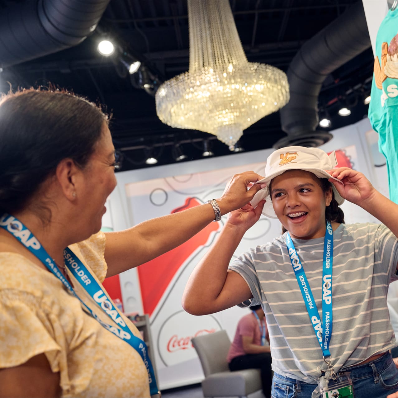 A mom and her two children are laughing and smiling while the daughter tries on a UOAP branded bucket hat. The family is shopping for exclusive UOAP merch within the AP Lounge located at Islands of Adventure.