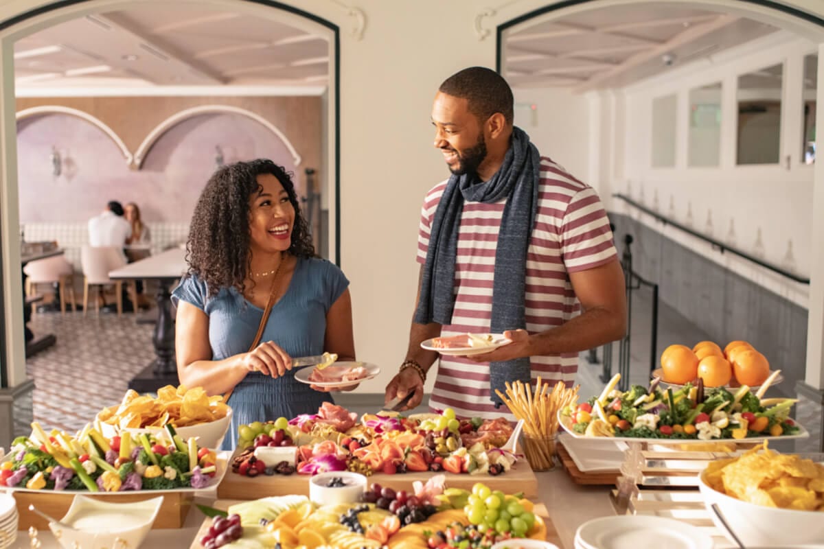 A smiling couple fill their plates with meats and cheeses from a decadent charcuterie table.