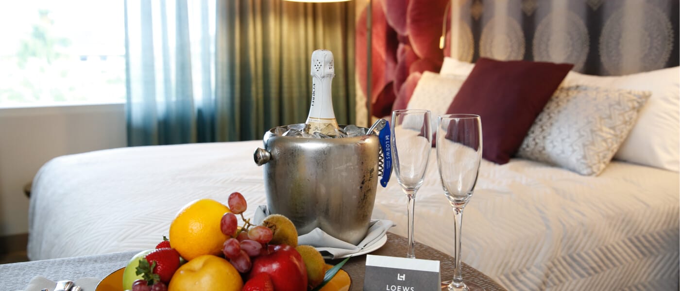 A plate of fresh fruit along with a bottle of champaign and glasses from room service is on display in front of the bed in a Guest Room in Loews Royal Pacific Resort in Universal Orlando.