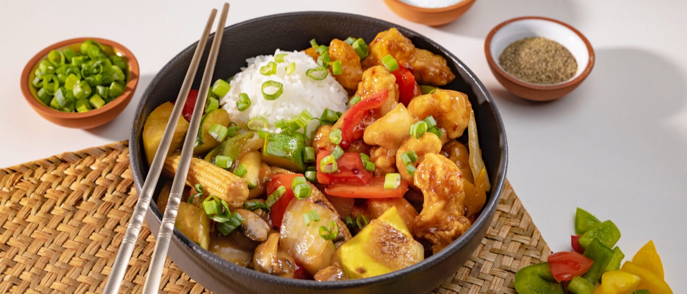 A bowl of crispy chicken, white rice, and vegetables on top of a bamboo placemat with metal chopsticks.