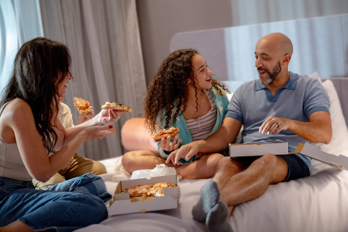A happy family of three laughing and eating pizza in their hotel room at Stella Nova Resort.