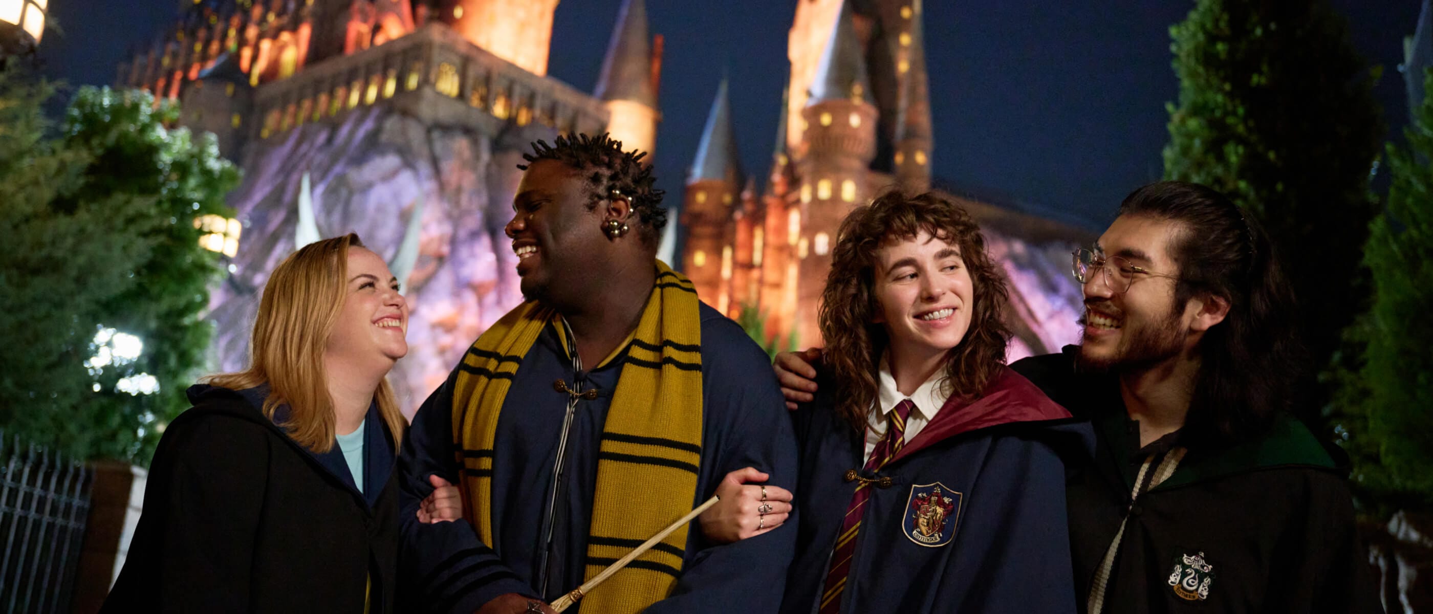Four friends with wizarding robes and accessories smiling and laughing together in front of Hogwarts™ castle lit up at night.