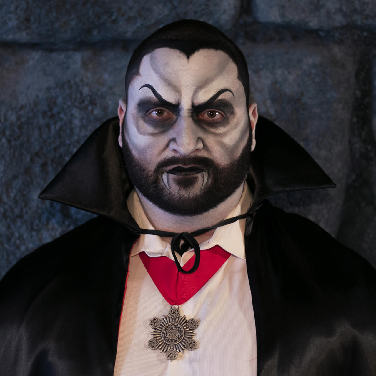 A guest is shown in a before and after shot, smiling on the left and transformed with makeup into a vampire with a cape on the right.