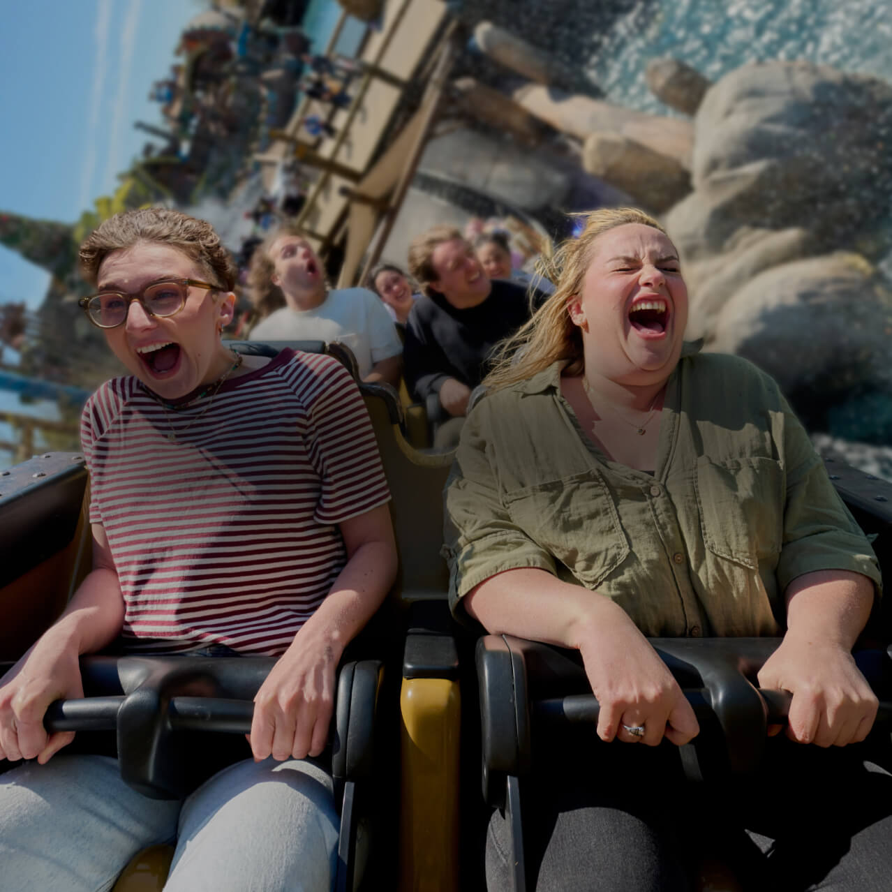 Two friends screaming and laughing as their roller coaster vehicle skims over rocks and water.