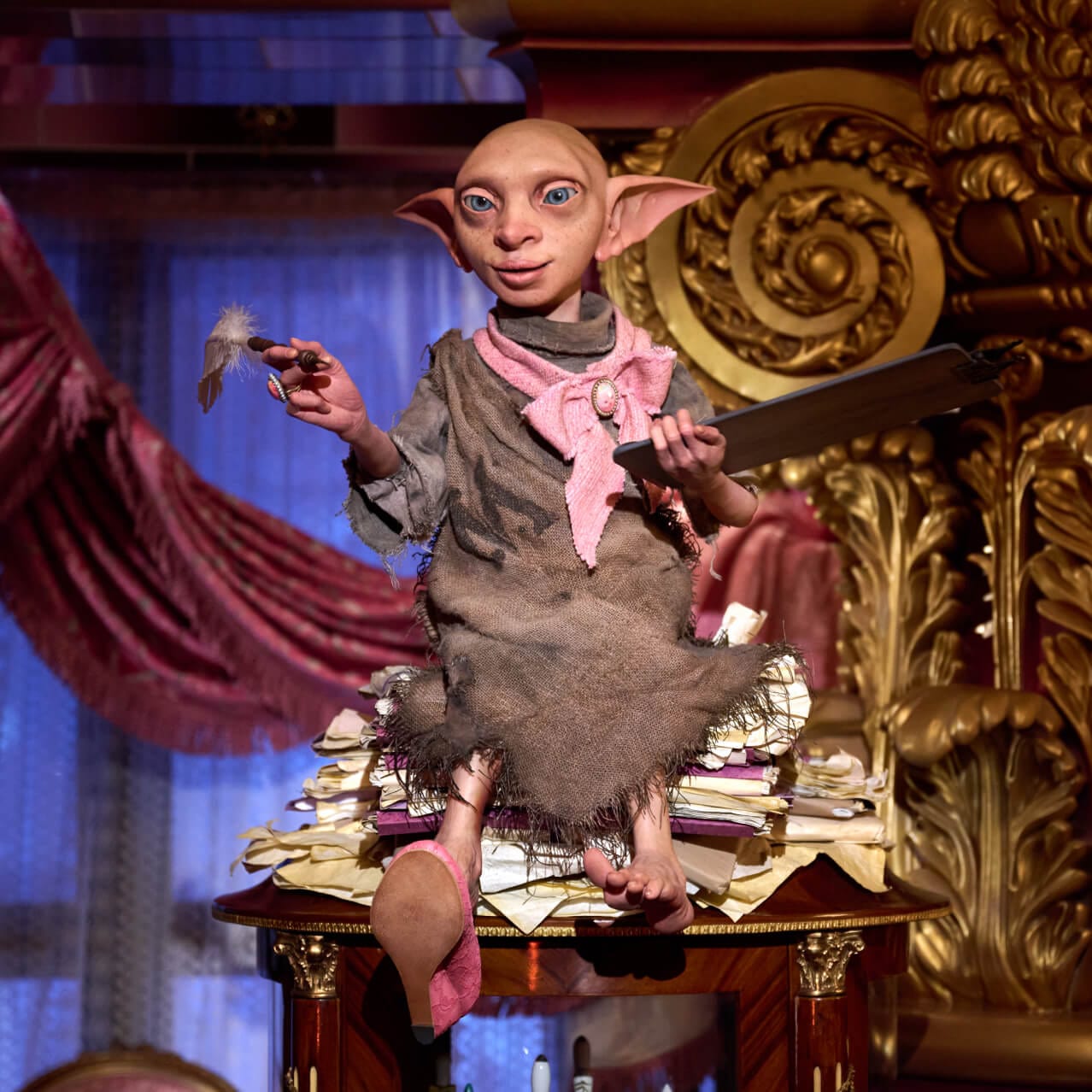 Higgeldy, a house elf, sits atop a pile of papers and she holds a quill and tablet.