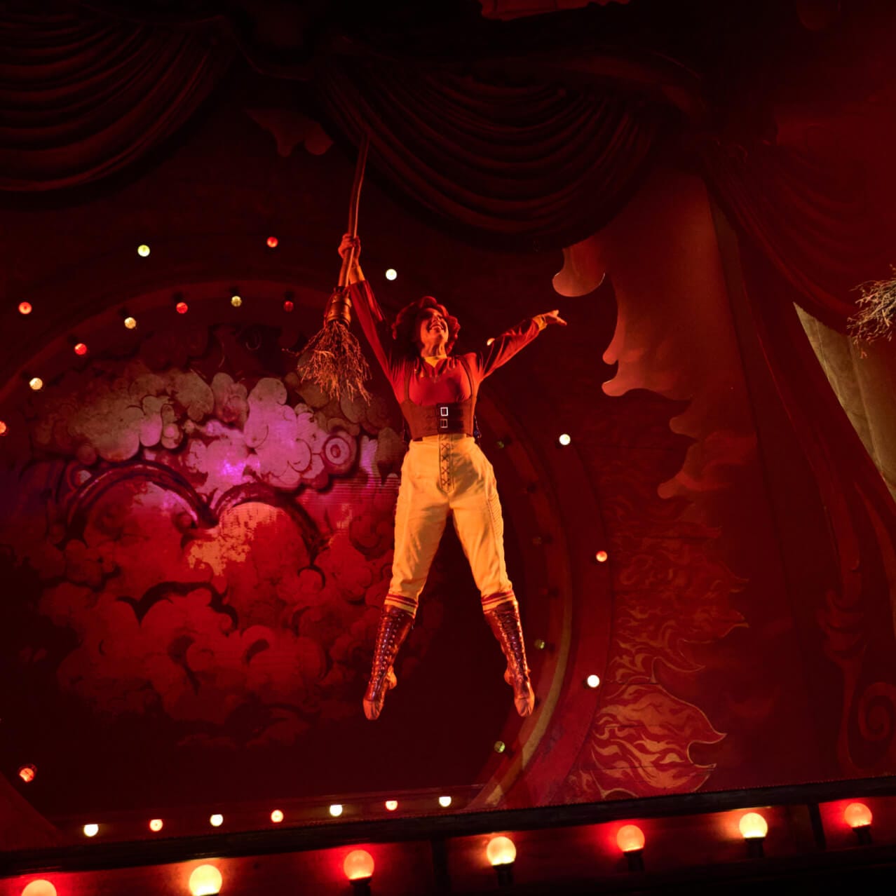 A witch floats into the air while two other witches hold their wands up, while performing on stage at Le Cirque Arcanus.